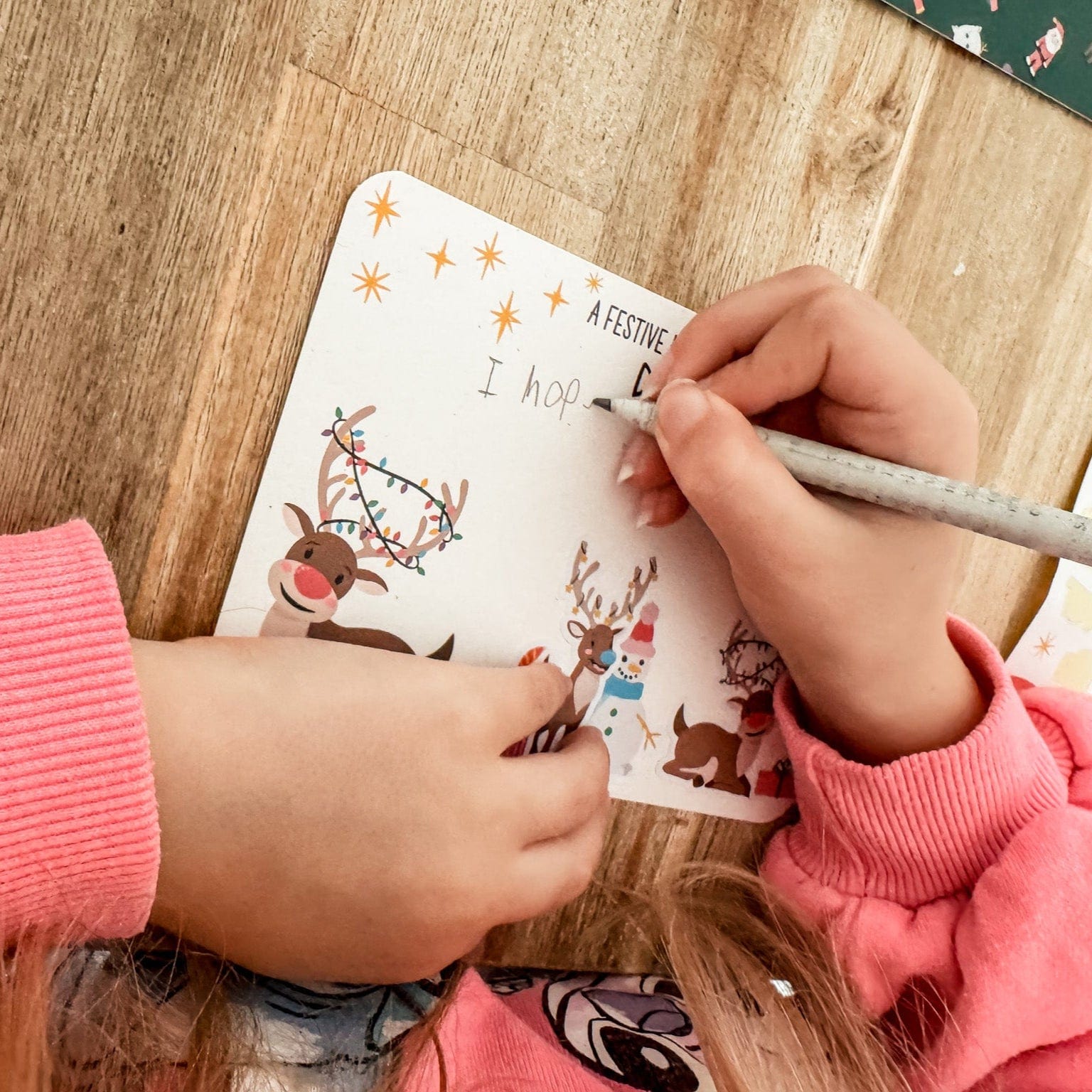 Child's hands writing a Christmas-themed postcard with a pencil on a wooden table.