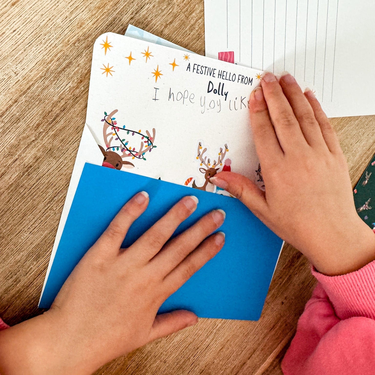 A child's hand closing a Christmas postcard with a reindeer illustration on a wooden table.