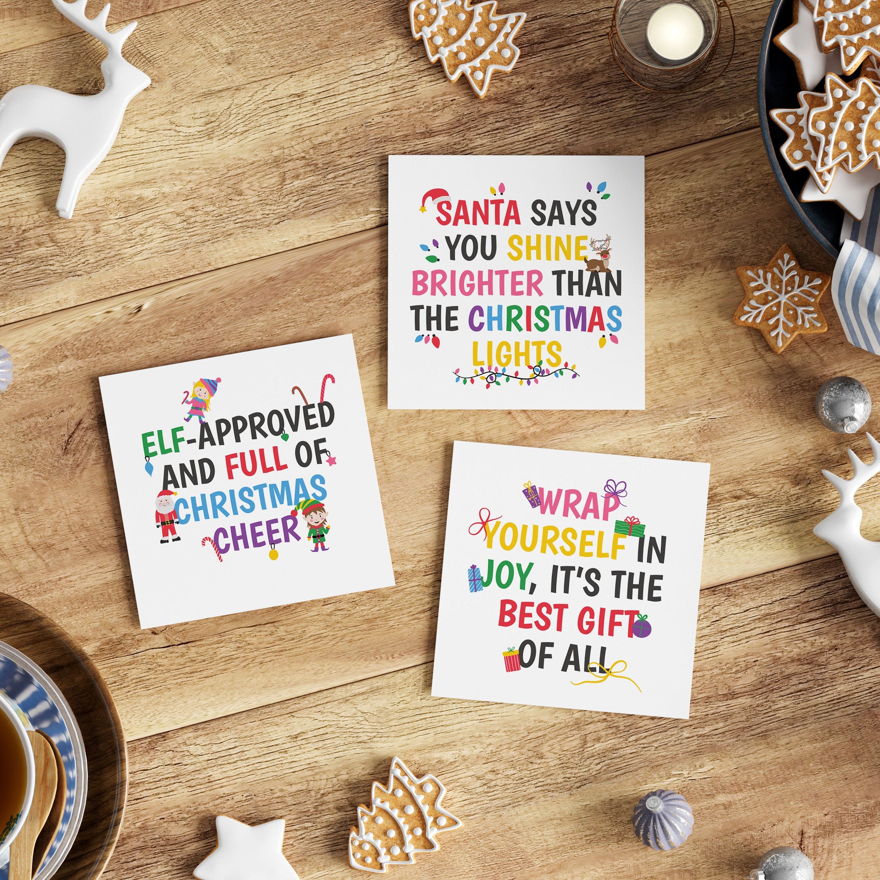 Three Christmas-themed cards on a wooden table with cookies and a cup of tea.