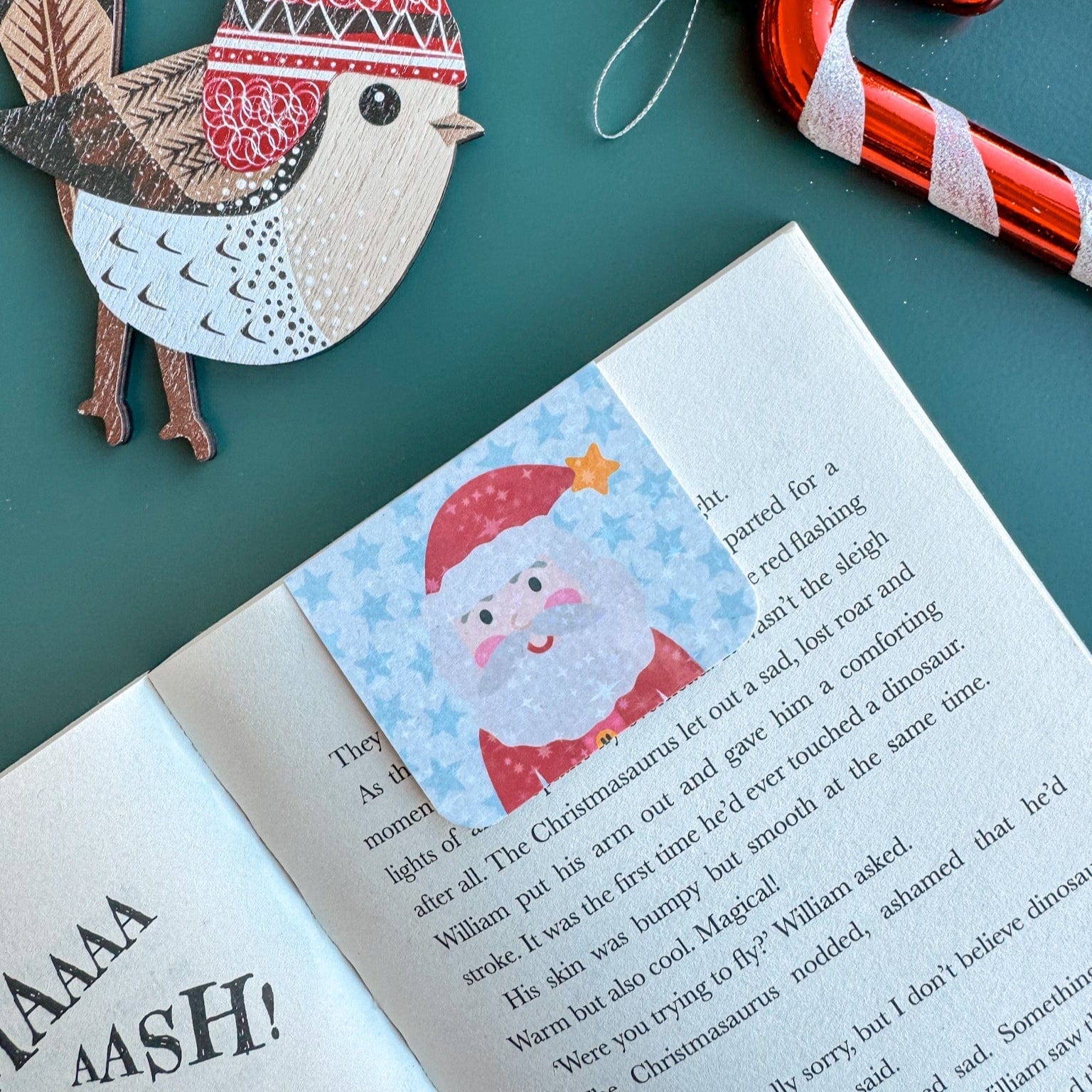 Open book with a Santa Claus bookmark, wooden bird ornament, and candy cane on a green background