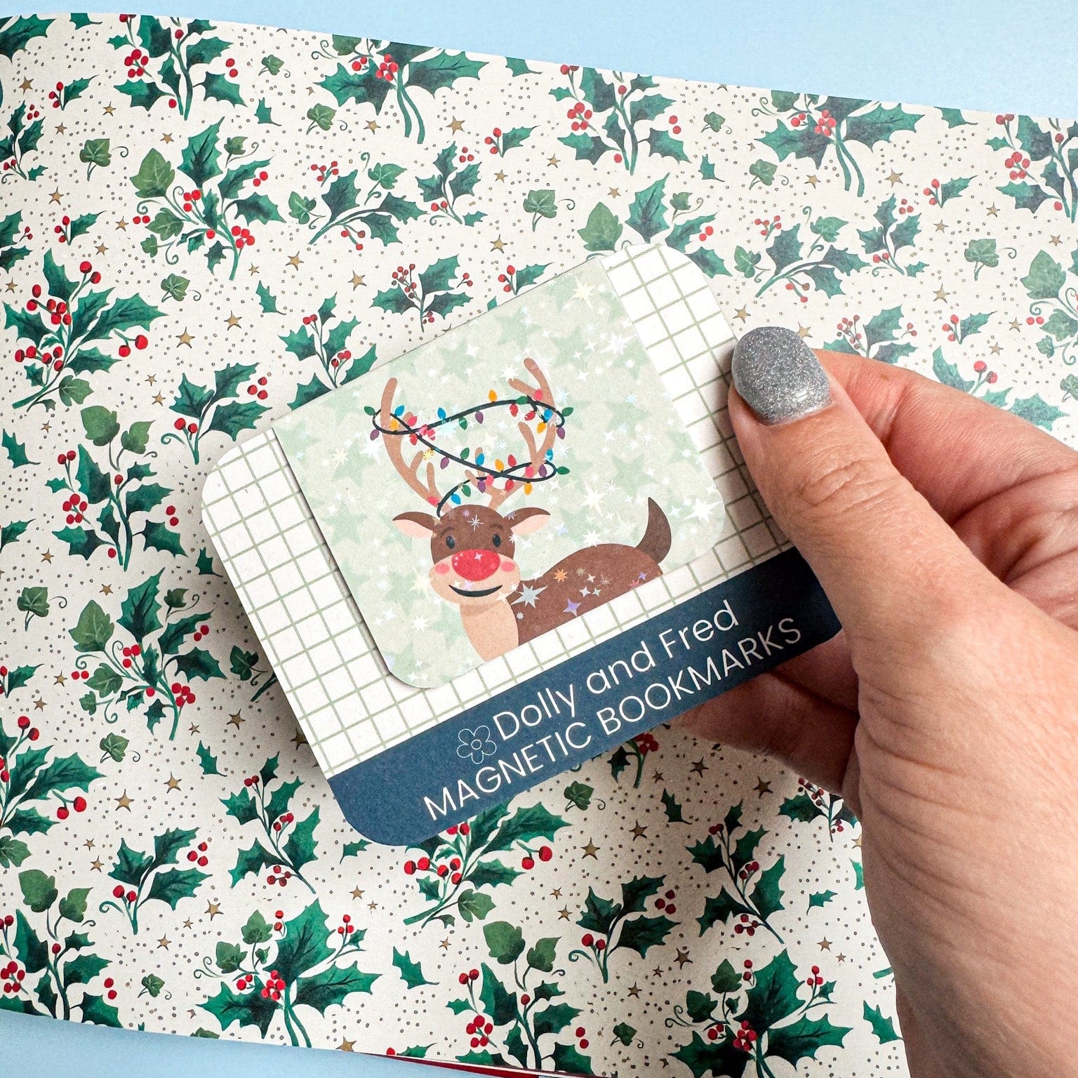 Hand holding a pack of Dolly and Fred magnetic bookmarks with a reindeer design on a holly leaf patterned surface.