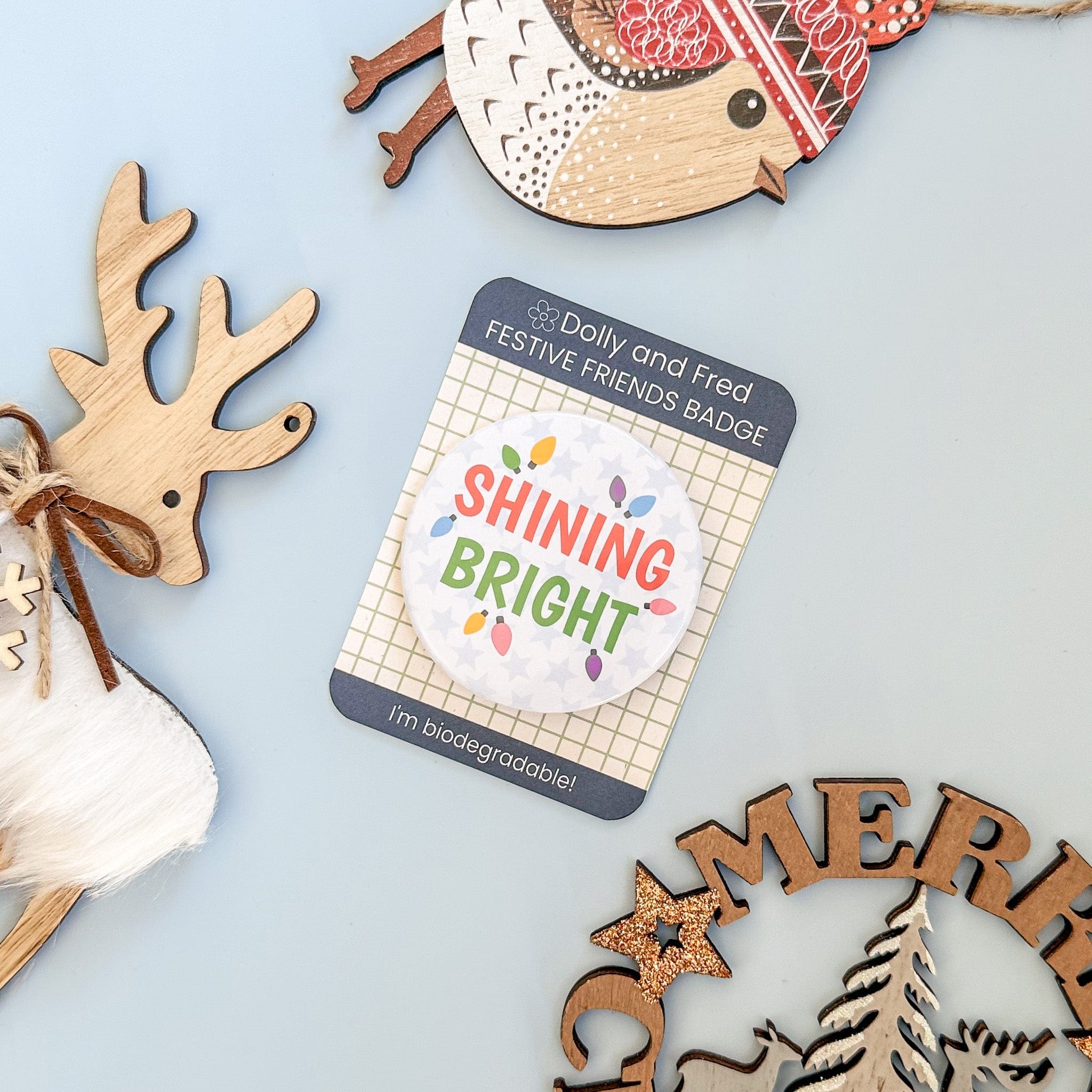 Festive badge with 'Shining Bright' text on a card, surrounded by wooden reindeer decorations on a light blue background.