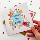 Child holding personalised teacher card with blue flowers and thank you message.
