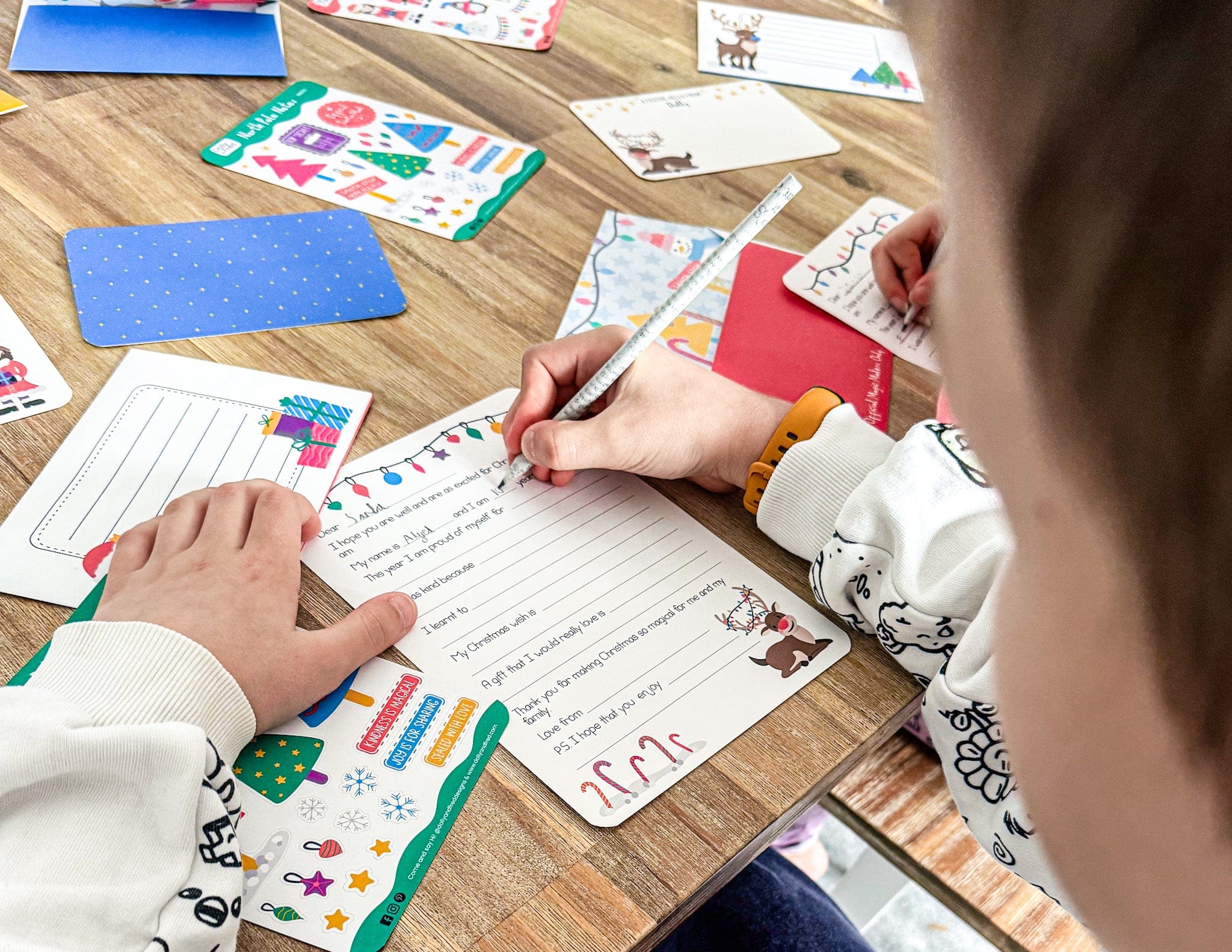 Children writing a letter ot santa surrounded by stickers and other stationery items at a wooden table