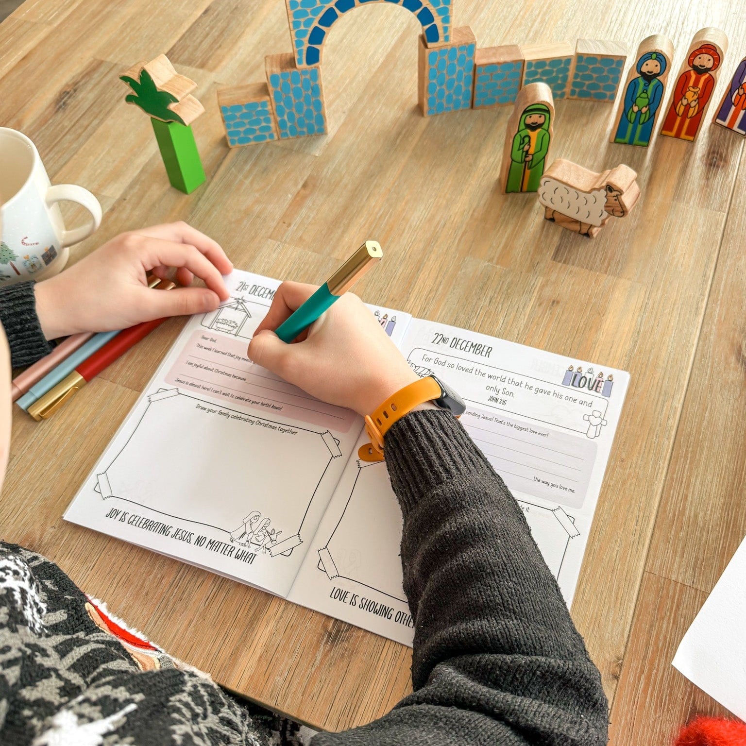 A boy colouring a picture with a crayon on a wooden table with toys and a mug.