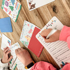 Children writing on stationery items with colourful Christmas designs on a wooden table.