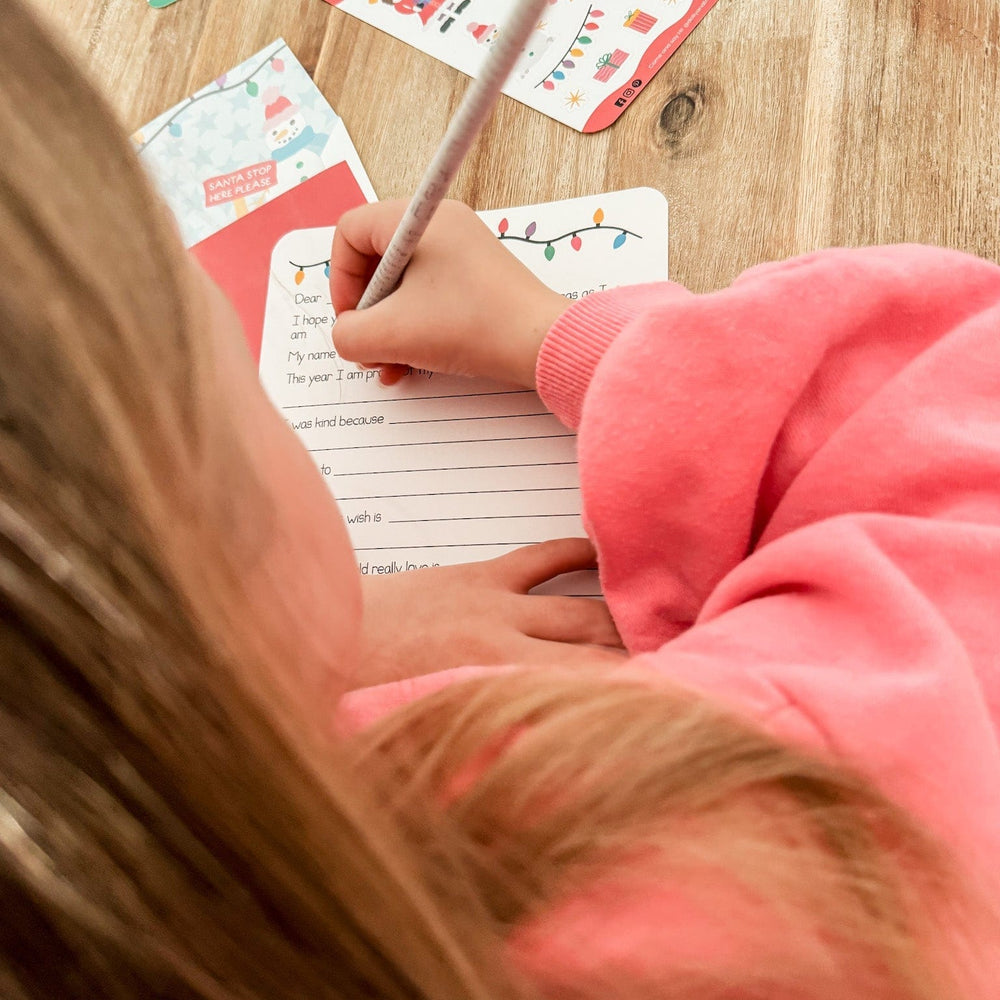 A child writing a letter to Santa with Christmas-themed cards on a wooden table