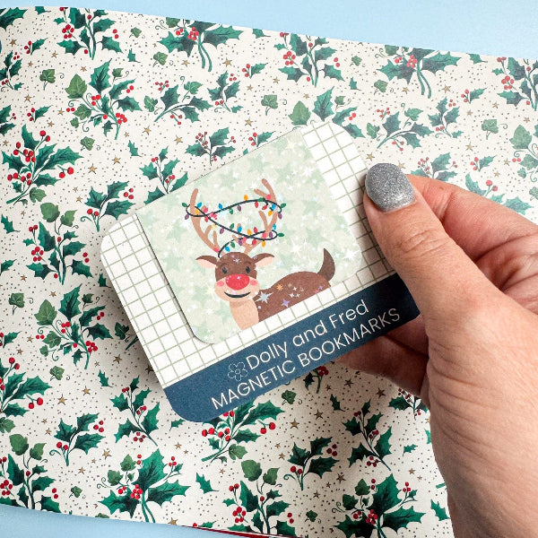 Hand holding a pack of Dolly and Fred magnetic bookmarks with a reindeer design on a holly leaf patterned surface.