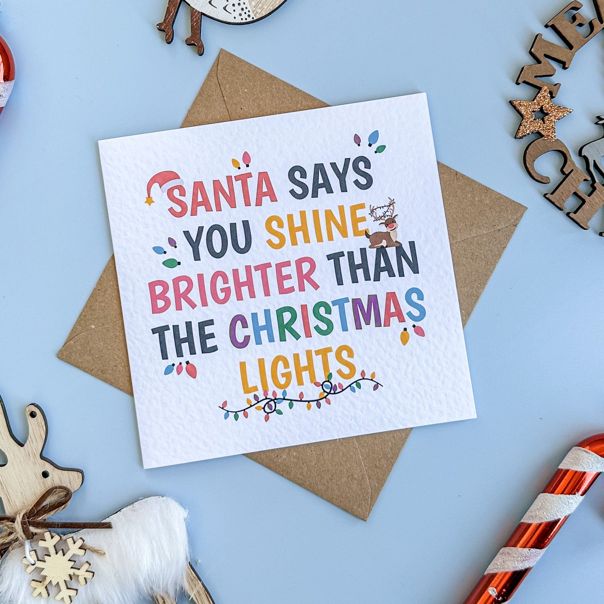 Christmas card with text 'Santa says you shine brighter than the Christmas lights' on a blue background with decorative items.