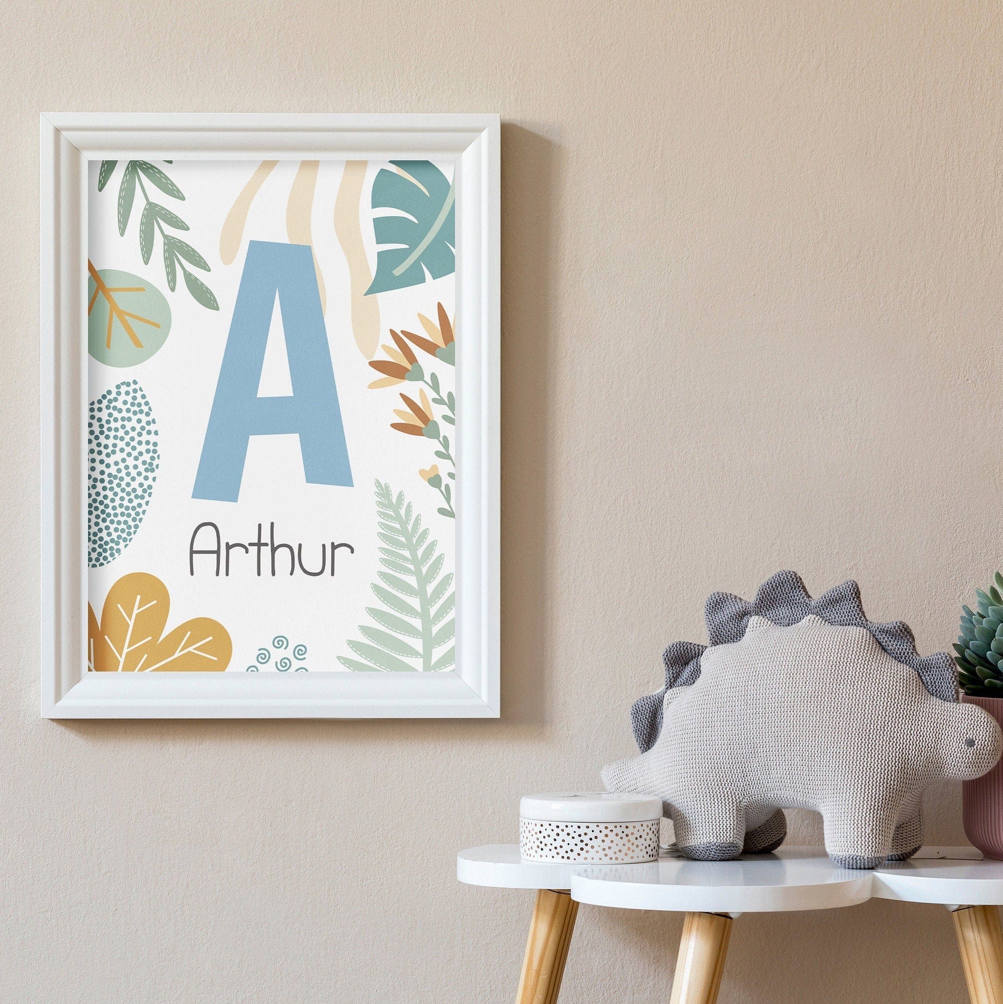 Framed artwork with a letter 'A' and name 'Arthur', dinosaur toy, and small plant on a table against a beige wall.