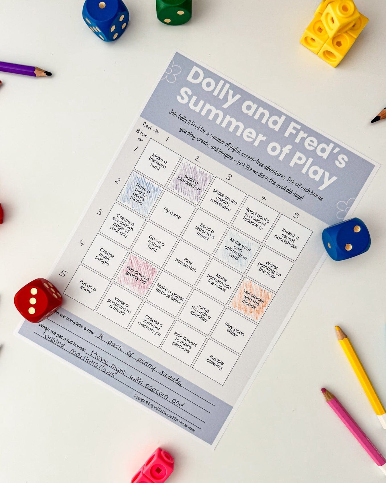 Flat lay of Dolly and Fred’s Summer of Play bingo card surrounded by colourful pencils and dice