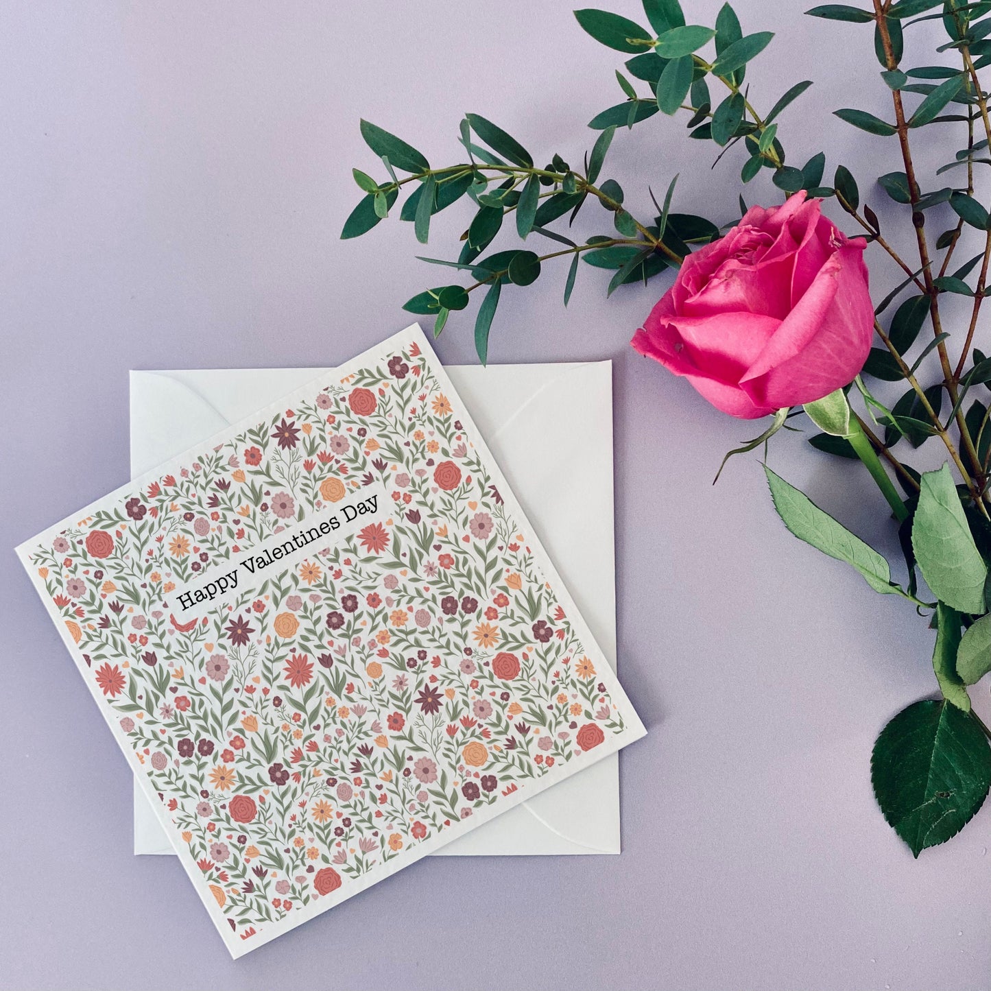 Square white card with a floral pattern in peach and pinks over it. There is the words happy valentines day towards the top of the card in a typewriter font with a white background.